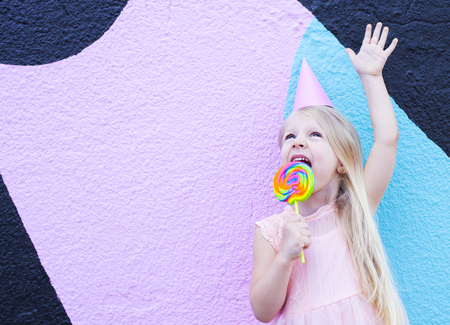 Cheerful little girl with birthday cap on head holding lollipop