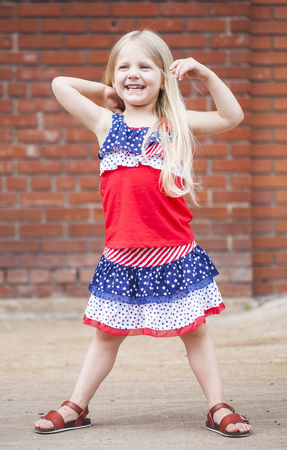 Portrait of laughing little girl touching hair outdoors. Cute blond girl wearing stars and stripes dress. Independence dayの写真素材