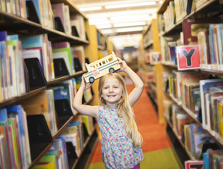 Cheerful schoolgirl holding toy school bus above her head in the libraryの写真素材