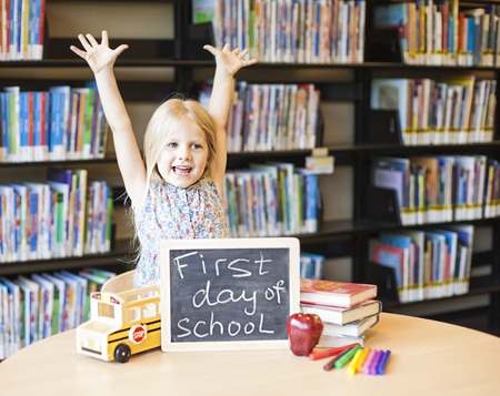 Cheerful girl raising hands at the table with chalkboard, books, markers and apple on it. First day of schoolの写真素材