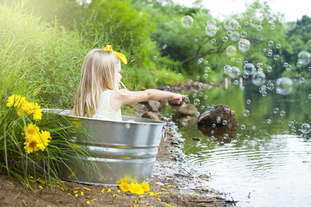 Cheerful little girl in metal bathtub at river playing with bubbles outside at summer timeの写真素材