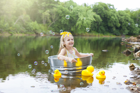 Cheerful little girl in metal bathtub with toy yellow ducks next to it and playing with bubblesの写真素材