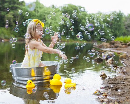 Cheerful little girl in metal bathtub with toy yellow ducks next to it and playing with bubblesの写真素材