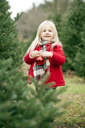 Portrait of happy little girl standing with bauble among fir trees. Girl hanging baubles on Christmas trees at farmの写真素材