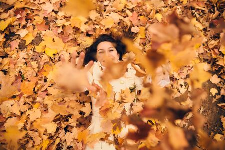 Happy young woman lying in foliage and throwing autumn leaves. Cheerful girl having fun in autumn park. Happiness, emotion, seasonの写真素材