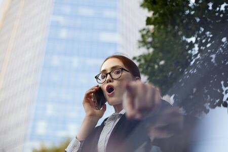 Excited businesswoman talking on mobile phone and pointing at camera. Portrait of female manager wearing glasses using smartphone pointing at youの写真素材