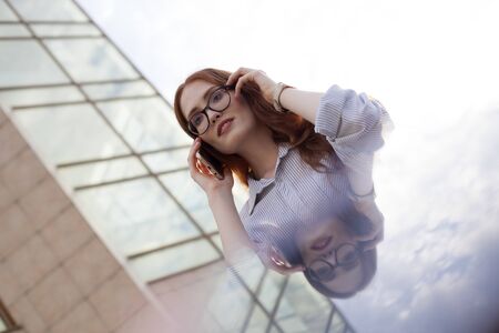 Concentrated young businesswoman talking on mobile phone. Portrait of redhead woman calling client at office buildingの写真素材