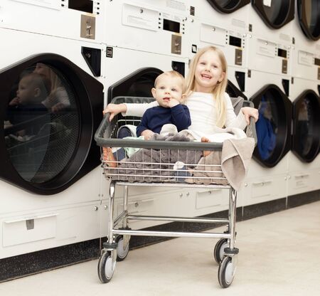 Portrait of little girl with her baby brother at laundry. Siblings sitting in cart with linen at washing machines. Family, housework, hygieneの写真素材