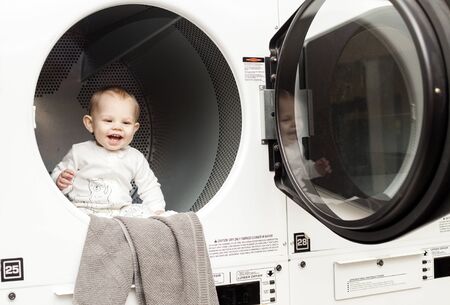 Portrait of laughing baby boy sitting in washing machine. Happy little toddler at laundry. Childhood...