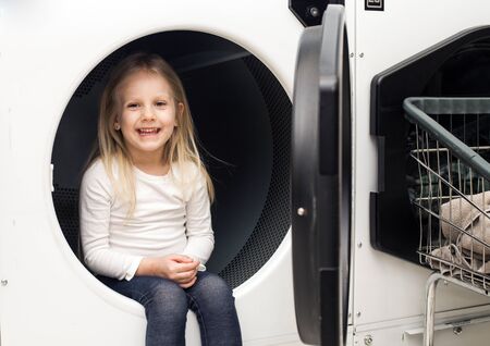 Portrait of cheerful little girl sitting in washing machine. Happy kid posing at laundry. Childhood and houseworkの写真素材