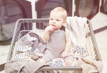 Portrait of baby boy sitting in cart among clothing at laundry. Serious little boy sucking his...