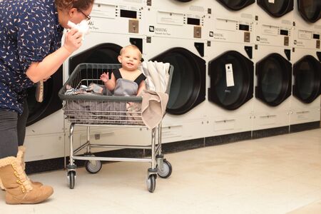 Mother with little son in cart putting linen in washing machine. Mid adult woman and her baby boy at laundry. Service and hygieneの写真素材
