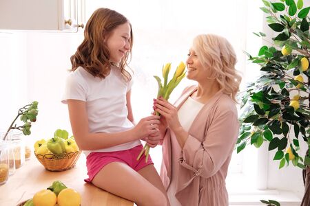 Smiling mother receiving flowers from teenage daughter. Girl sitting on table and congratulating mom with birthday or Mother s Day at kitchen. Holiday, family, springtimeの写真素材