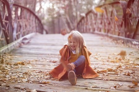 Portrait of excited little girl sitting on bridge in autumn. Playful child wearing coat walking in fall. Childhood, season, weekendの写真素材