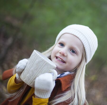 Portrait of happy little girl drinking tea outdoors. Face of cute child wearing knitted hat holding cup. Childhood, season, hot drinkの写真素材