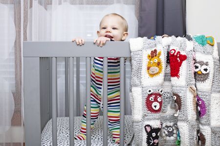 Portrait of happy baby boy standing on crib. Little kid wearing jumpsuit in nursery. Childhood,...