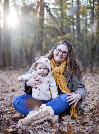 Portrait of happy young mother posing with baby son in autumn wood. Woman wearing glasses and coat sitting on foliage with little son, looking at camera and smiling. Motherhood, maternity leaveの写真素材
