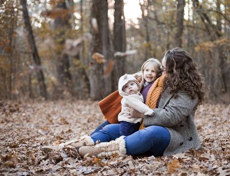 Portrait of happy mother sitting with two children on foliage. Young woman wearing coat and glasses walking with baby son and little daughter in autumn wood. Motherhood, family, weekendの写真素材