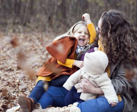 Cheerful little girl sitting with mother and brother on foliage. Young woman walking with her little daughter and baby son in autumn wood. Motherhood, family, weekendの写真素材