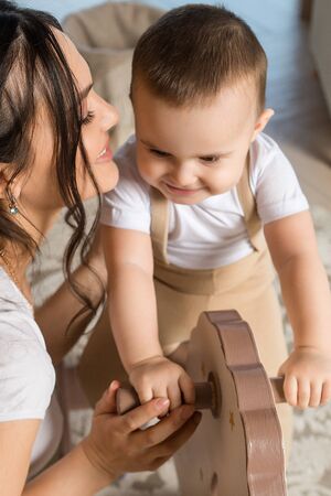 Happy young mother kissing her little son in nursery. Smiling baby boy riding toy in playroom. Childcare, family, motherhoodの写真素材