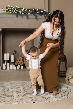 Happy young mother teaching baby son to walk at home. Portrait of little boy making first steps with his mom in living room. Baby steps, childcare, motherhoodの写真素材