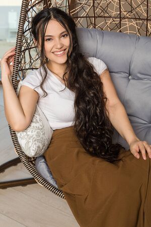 Portrait of smiling beautiful woman in hanging wicker chair. Pretty young lady wearing white top and long brown skirt posing in studio. Natural beauty, South Eastern womanの写真素材