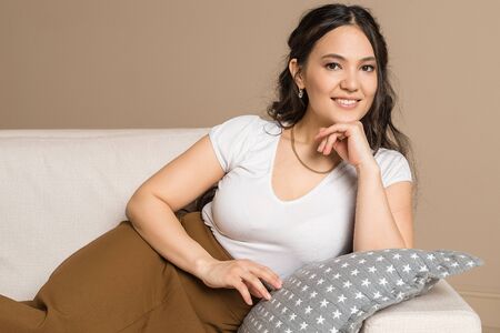 Portrait of happy beautiful woman resting on coach. Young girl posing with hand on chin, looking at camera and smiling. Natural beauty, South Eastern womanの写真素材