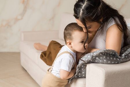 Happy young mother resting with baby son on couch. Portrait of brunette woman lying with child. Childcare, family, motherhoodの写真素材