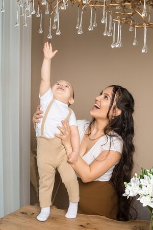 Portrait of happy Portrait of happy baby boy playing with mother at home. Little son standing on table and raising hand to touch glass pendants on chandelier. Childcare, family, motherhoodの写真素材