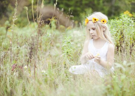 Portrait of serious little girl sitting in grass outdoors. Girl wearing white dress and wreath walking in summer meadowの写真素材