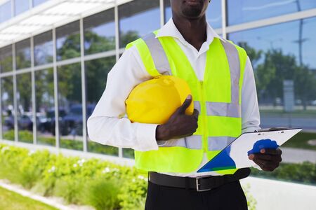 Close-up of engineer with hardhat and clipboard. African American contractor standing at office buildingの写真素材
