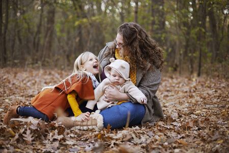 Portrait of young mother resting with her children in wood. Woman sitting on foliage with baby son and little daughter and laughing. Motherhood, young mother, childcareの写真素材