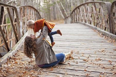 Happy mother playing with little daughter on bridge. Young woman sitting on bridge in foliage and...