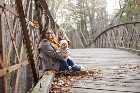 Portrait of happy mother sitting on bridge with children. Young woman walking with baby boy and little girl in autumn. Family, weekend, seasonの写真素材