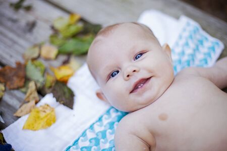 Close-up of smiling baby son lying on towel among leaves. Portrait of little boy looking at camera and smiling. Newborn child conceptの写真素材