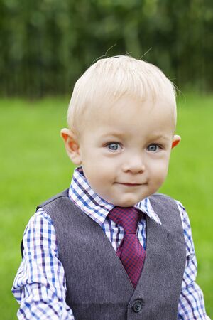 Portrait of smiling little boy wearing formal suit looking at camera outdoors. Toddler wearing white checked shirt, waistcoat and necktie. Childs fashion, little businessmanの写真素材