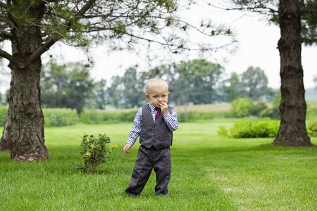 Portrait of serious little boy walking alone outdoors. Toddler wearing formal suit standing on lawn among trees. Little businessman ,childhoodの写真素材