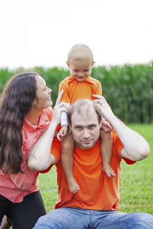 Portrait of happy family playing with son outdoors. Couple sitting together on grass, little boy sitting on neck of his father. Family, parenthood, relationshipsの写真素材