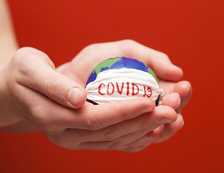 Stop corona virus. Conceptual photo of male hands holding globe with face mask on it isolated on red backgroundの写真素材