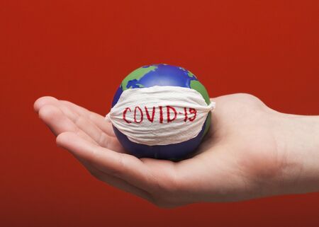 Stop corona virus. Conceptual photo of male hands holding globe with face mask on it isolated on red backgroundの写真素材