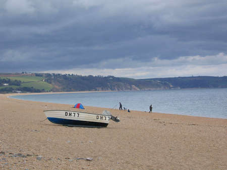 Small fishing boat moored on a beach in South Devon in England with two men rod fishing in the distance.の写真素材