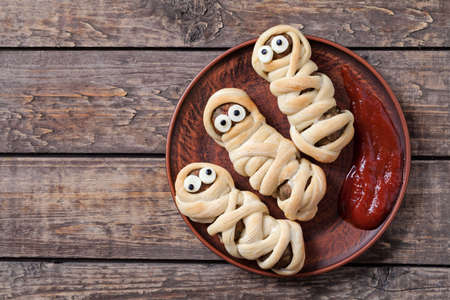 Mummy sausages scary halloween party food decoration wrapped in dough with blood sauce on clay dish. Vintage wooden backgroundの写真素材