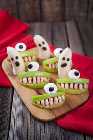 Scary halloween food edible monsters healthy celebration party decoration recipe. Crazy cyclop apple mouth with teeth and banana ghosts, red fabric on vintage wooden backgroundの写真素材