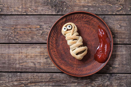 Funny halloween sausage mummy wrapped in dough celebration food on vintage wooden background. Rustic style and natural light.の写真素材
