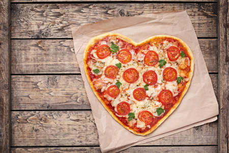 Heart shaped pizza margherita with tomatoes and mozzarella for Valentines Day on vintage wooden background. Food concept of romantic love. Rustic style, top view.の写真素材
