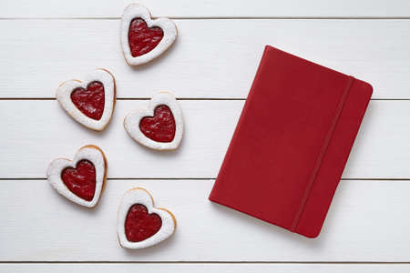 Heart shaped cookies with empty red notebook, composition on white wooden background for Valentines day. Food concept of romantic love. Top view, flat lay.の写真素材