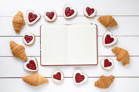 Empty notebook frame for design text, croissants and heart shaped cookies on white wooden background. Valentines day food concept of romantic love. Top view, flat lay.の写真素材