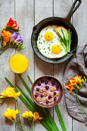 Concept of breakfast with bright spring mood, oatmeal porridge with nuts, blackberry, juice in glass and fried eggs in cast iron pan on vintage wooden table . Top view.の写真素材
