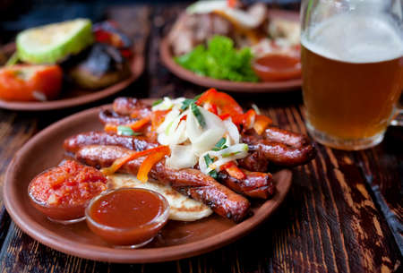 Barbecued pork or lamb meat sausages grill with salad, vegetables, glass of beer and sauce on clay dish. Vintage wooden table background.の写真素材