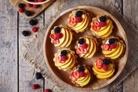 Tartlets with vanilla custard and fresh raspberries, blackberry and peach served on wooden tray with baking forms, kitchenware on the old wooden table. Top view. Dark rustic style.の写真素材